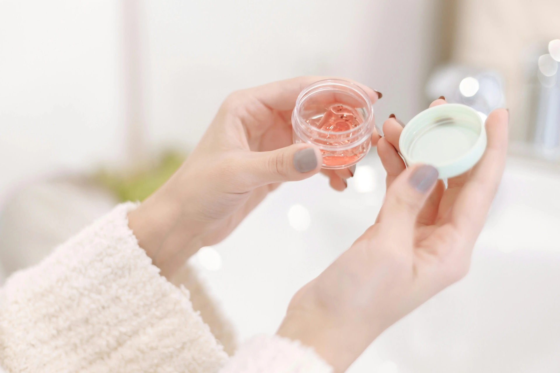 Manos sostienen frascos de vidrio con líquido rosado y crema blanca, en ambiente luminoso y desenfocado.