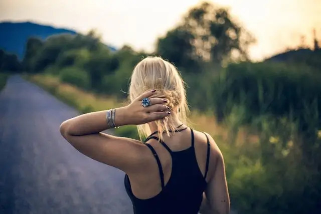 Mujer rubia con top negro camina por un camino rural, cubriéndose el rostro con la mano.