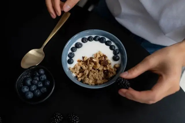Manos sirven un tazón con granola y arándanos, sobre mesa oscura con cucharas y frutas.