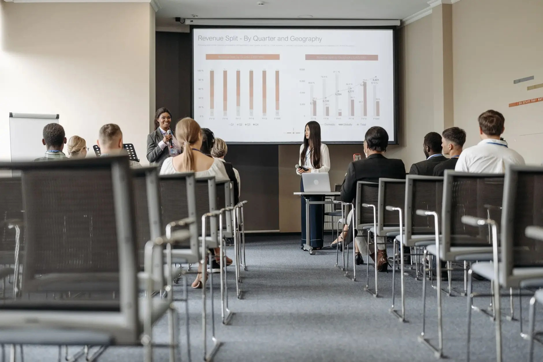Presentación empresarial en aula, ponente con micrófono y audiencia frente a gráficos en pantalla.
