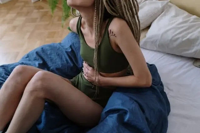 Mujer con top verde y vestido azul, sentada en cama, con tatuaje en el brazo y cabello trenzado.