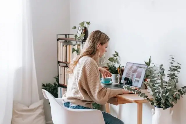 Mujer sentada en casa con laptop, taza y libros, rodeada de plantas y luz natural.