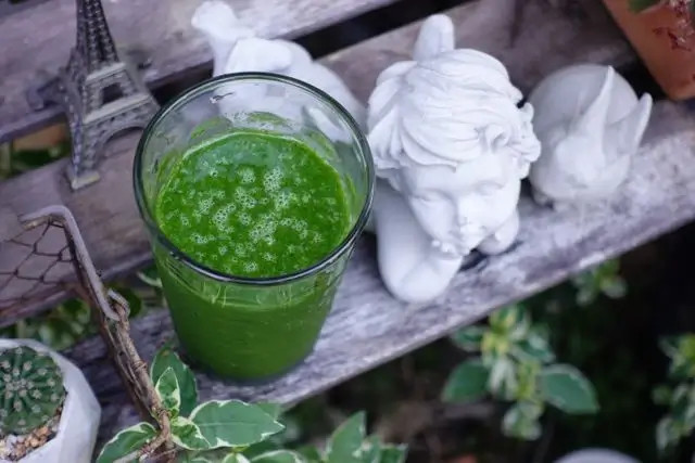 Vaso con jugo verde sobre bandeja de madera, junto a estatuillas blancas de ángeles.