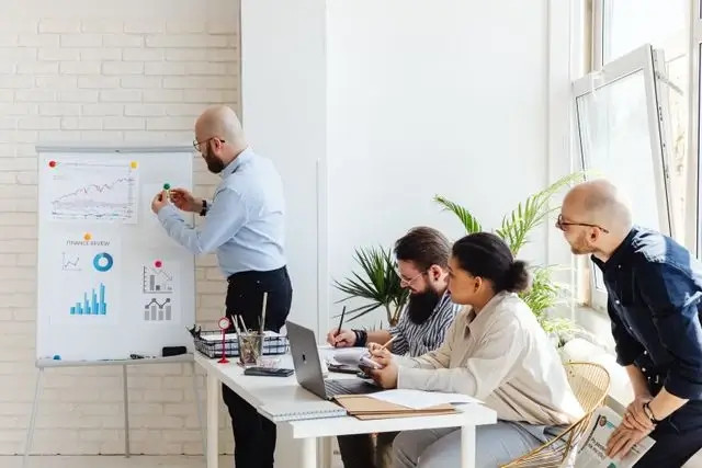 Hombre explicando gráficos en una pizarra blanca mientras un equipo trabaja en una mesa con laptops.