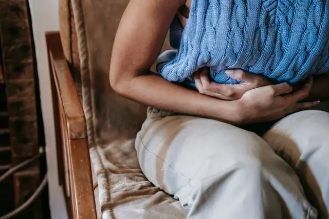 Persona con suéter azul sostiene el abdomen, sentada en una silla, expresión de dolor o malestar.
