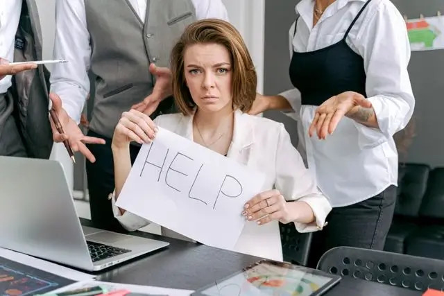 Mujer en oficina sostiene un cartel con “HELP” mientras compañeros a su alrededor gesticulan preocupados.