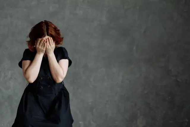 Mujer con vestido negro se tapa la cara con las manos, fondo gris oscuro, expresión de angustia.