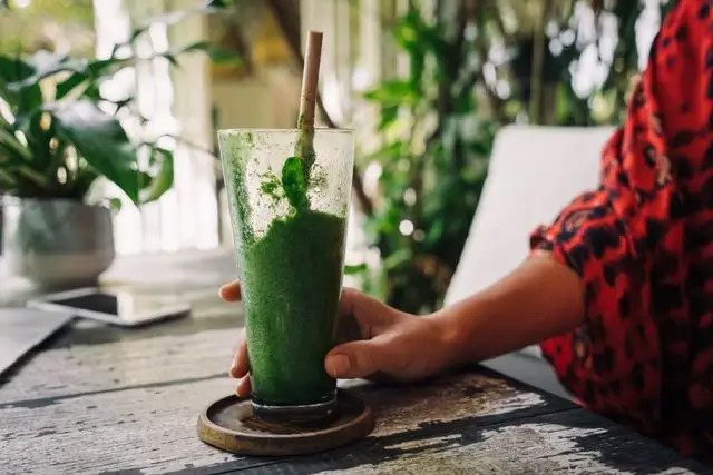 Persona sostiene un vaso alto con batido verde y pajita, sobre una mesa de madera al aire libre.