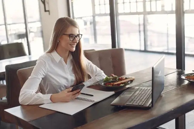 Mujer sonriente con gafas trabaja en oficina moderna, usando teléfono y computadora junto a un plato.