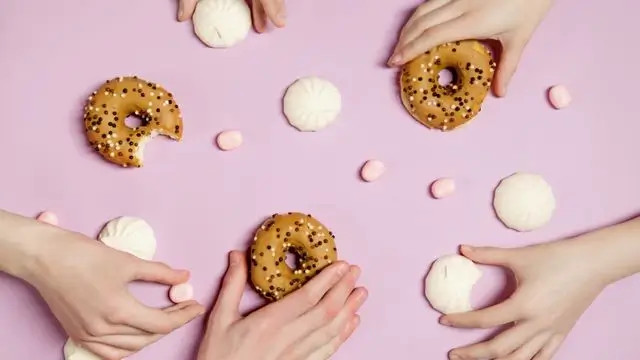 Manos decorando donas con chispas y crema blanca sobre fondo morado, con mini malvaviscos alrededor.