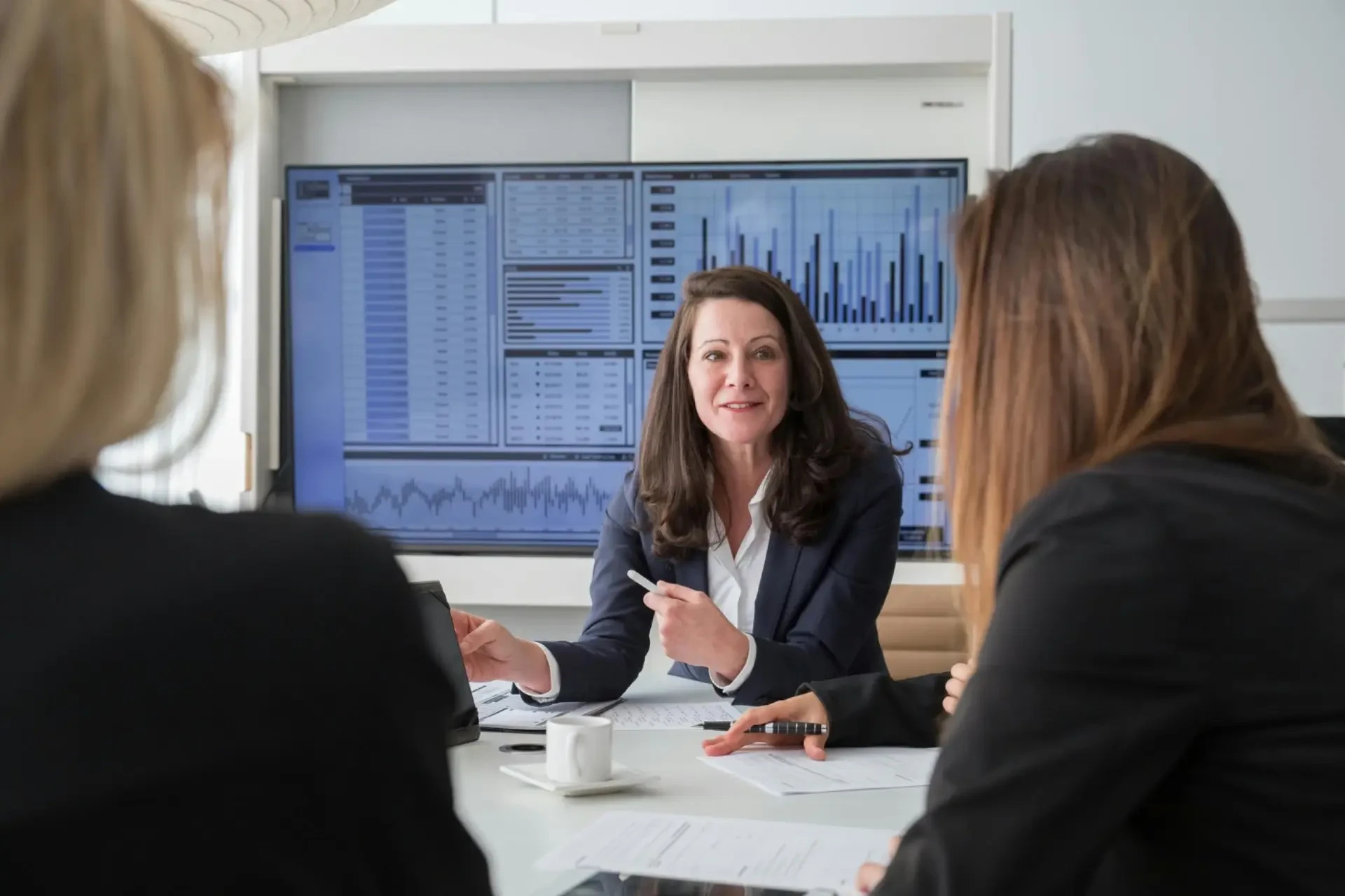 Ejecutiva conversa en una oficina con pantallas de gráficos y datos financieros detrás, durante una reunión.
