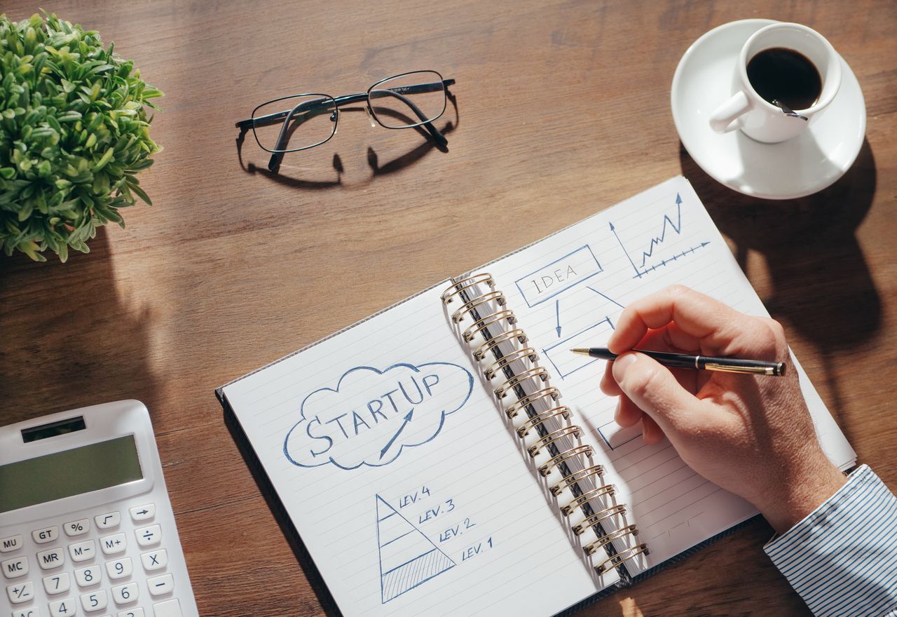 Mano escribe en cuaderno con gráfico “STARTUP”, calculadora, gafas y taza de café sobre mesa de madera.