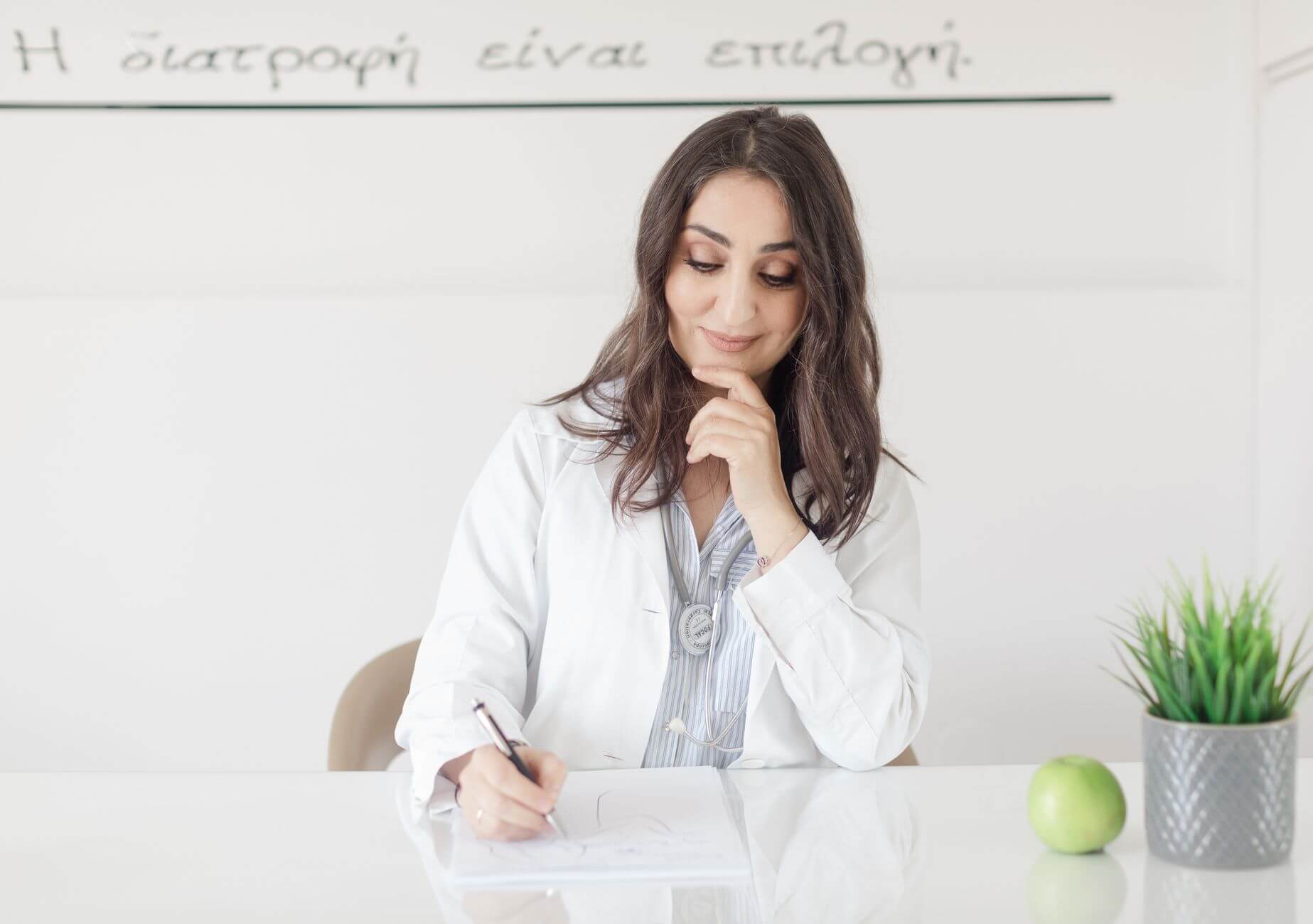 Mujer doctora con bata blanca, estetoscopio y bolígrafo, sentada frente a pizarra con texto. Nutricionista.