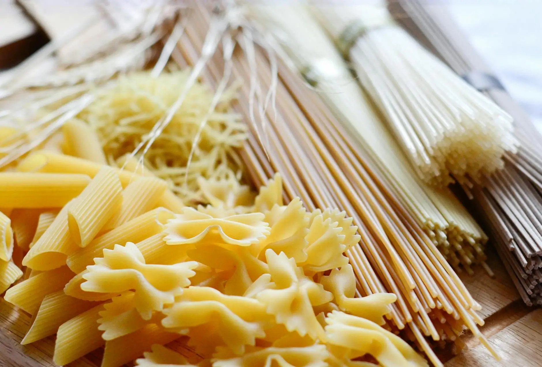 Pasta penne amarilla y espaguetis sobre tabla de madera, con tenedores de metal al fondo desenfocado. Carbohidratos.