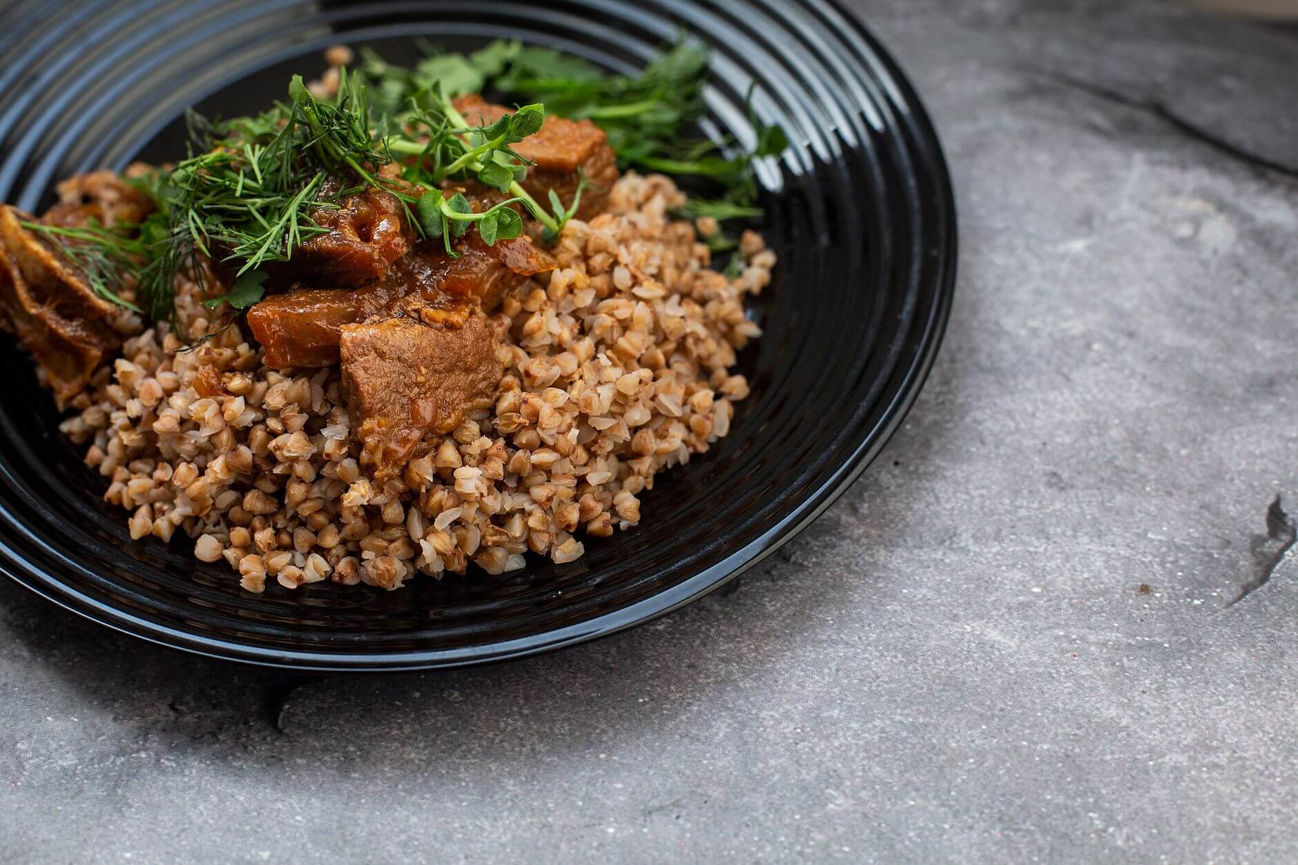 Plato negro con trigo sarraceno, carne guisada y hierbas frescas, sobre superficie gris. Carbohidratos.